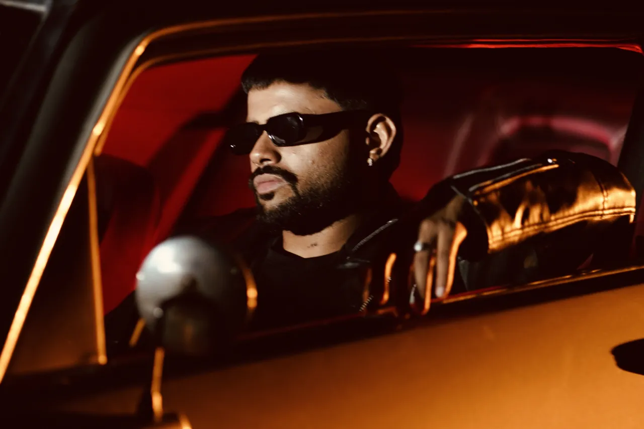 Badal in dark sunglasses behind the wheel of a car, lit in red and amber