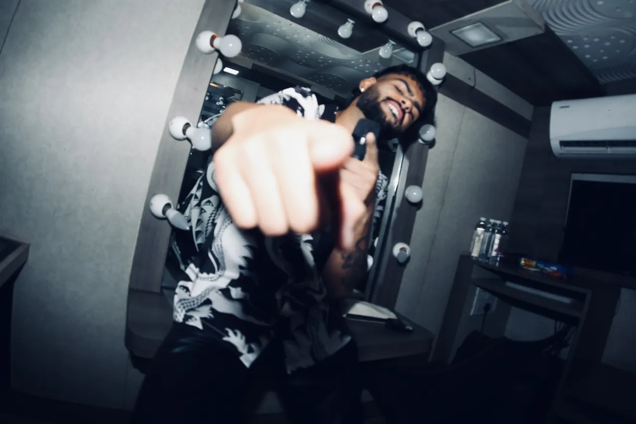 Badal pointing at the camera backstage, in front of a dressing-room mirror ringed with bulbs